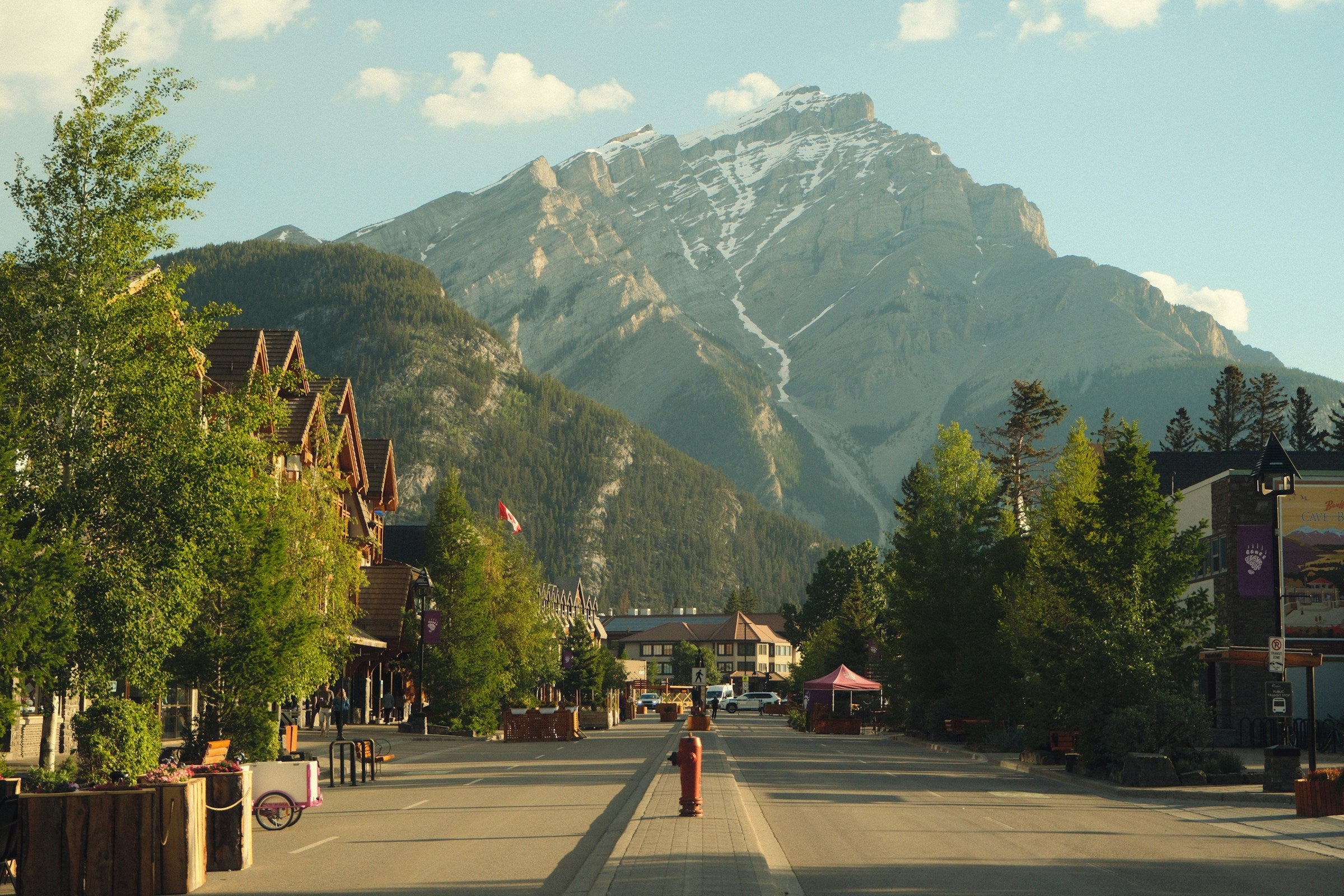 A scenic mountain town street view of Banff, Alberta with tall green trees, buildings along the sides, and a large snow-dusted mountain rising in the background under a blue sky.